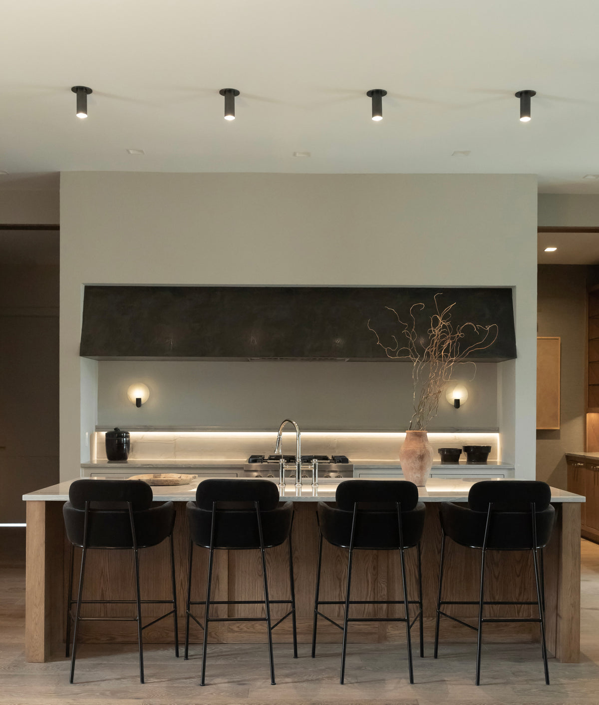 A modern kitchen with a large island featuring a marble countertop, four black barstools, a sink, and a decorative vase. Neutral tones prevail, complemented by under-cabinet lighting and Long Made Co.'s Brass Cylinder Flush Spot fixtures above the island.