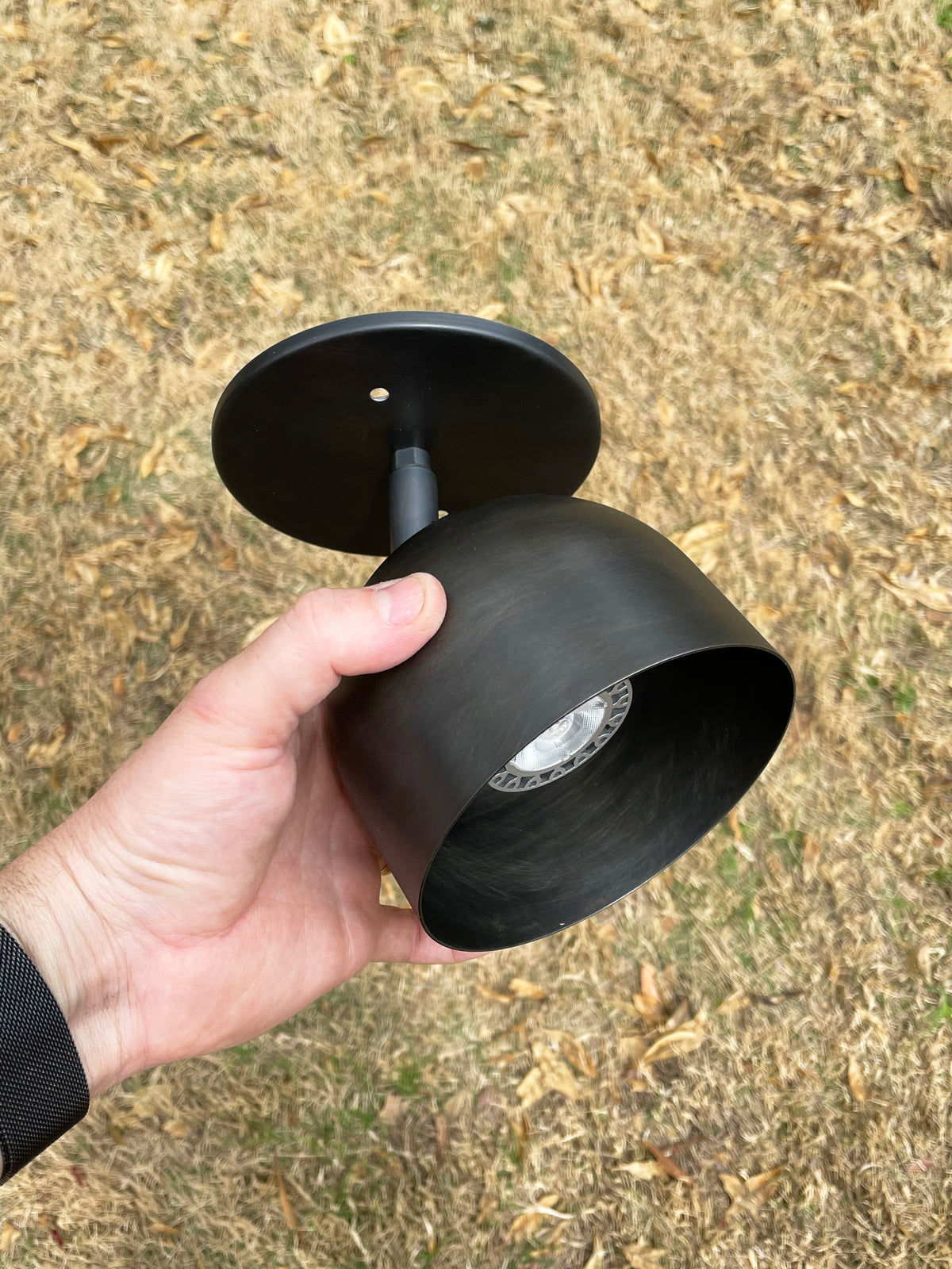 A hand holds the Long Made Co. Monaco Spot, a black outdoor downlight with a round base and cylindrical shade, made of solid steel, set against dry brown grass with scattered leaves.