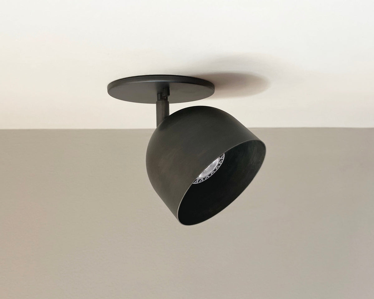 The Long Made Co. Monaco Spot is a black, modern ceiling-mounted downlight with solid steel construction, angled downward against a white ceiling and light gray wall.