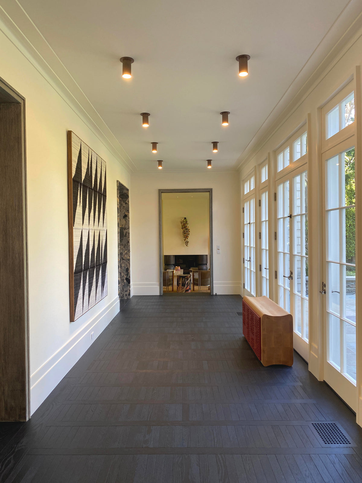 A bright hallway with dark wood floors, Long Made Co. Cove Flush Spot ceiling lights, large right-side windows, a geometric black-and-white wall hanging, and a brown bench beneath the windows. A dining area is visible through an open doorway.