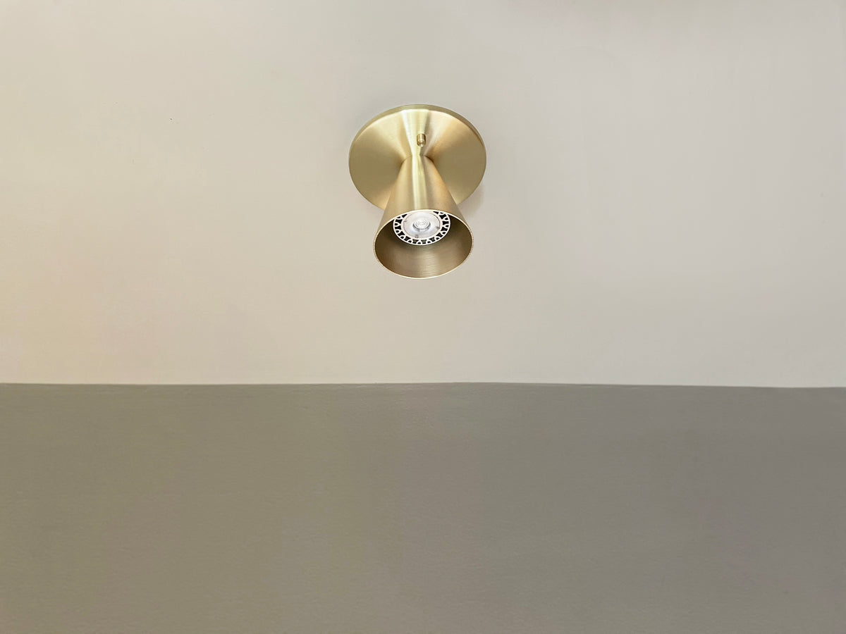 The Tiel Flush Spot by Long Made Co. is a modern solid brass ceiling fixture with a cone shade, mounted on a two-toned wall. It provides a stylish monopoint design and serves as a sleek alternative to recessed can lights.
