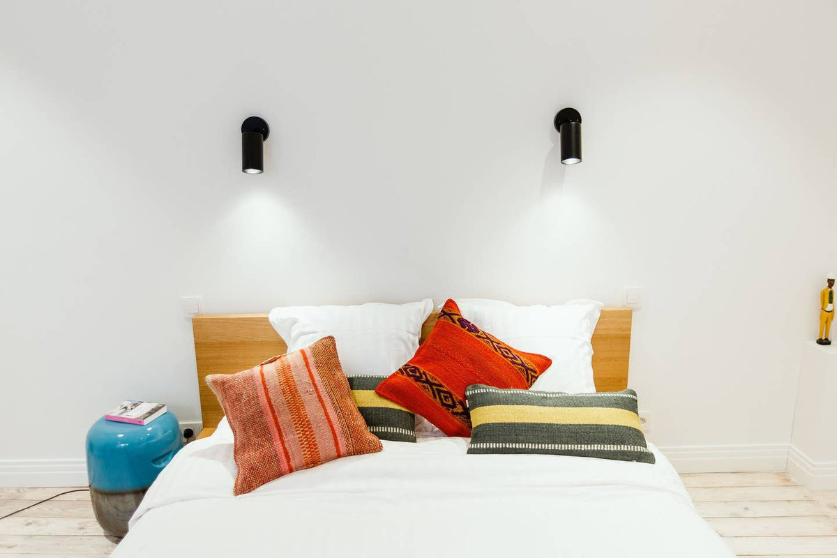 A neatly made bed with a wooden headboard, white bedding, and four colorful patterned pillows sits beneath black Cove Wall Lamps by Long Made Co. A blue side table with a book is placed beside the bed.