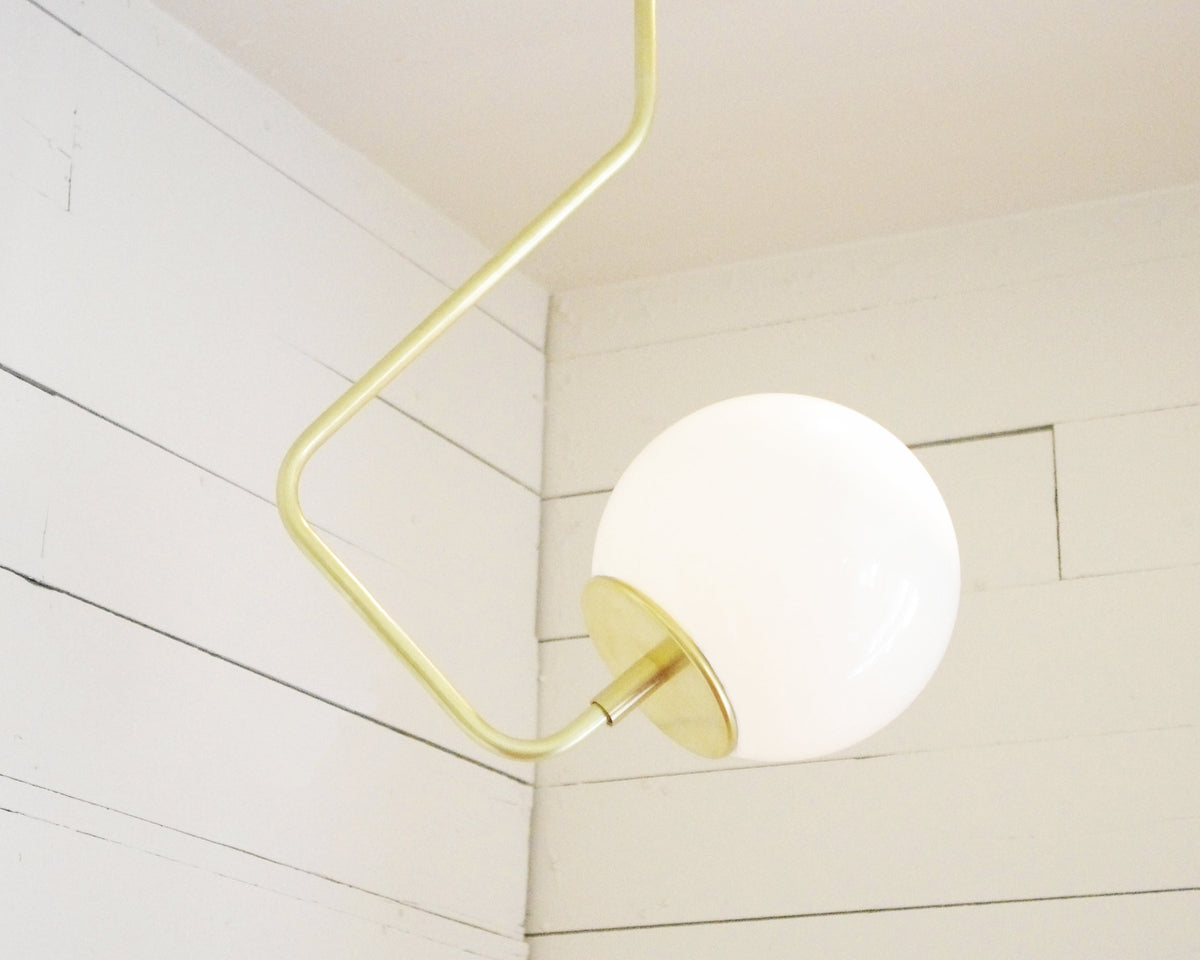 The Sphere Solo Bent Arm by Long Made Co., a modern handmade brass fixture with a hand-blown glass globe and gold angular arm, is mounted on a white wooden plank wall in the corner of a room.