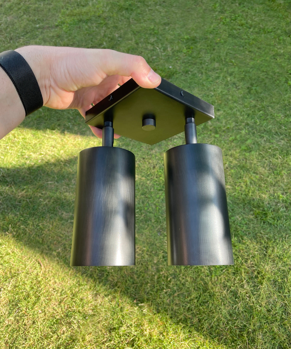 A hand holds the Long Made Co. Cove Double Spot, a black double-headed outdoor light fixture with solid steel spotlights, above a grassy lawn on a sunny day.