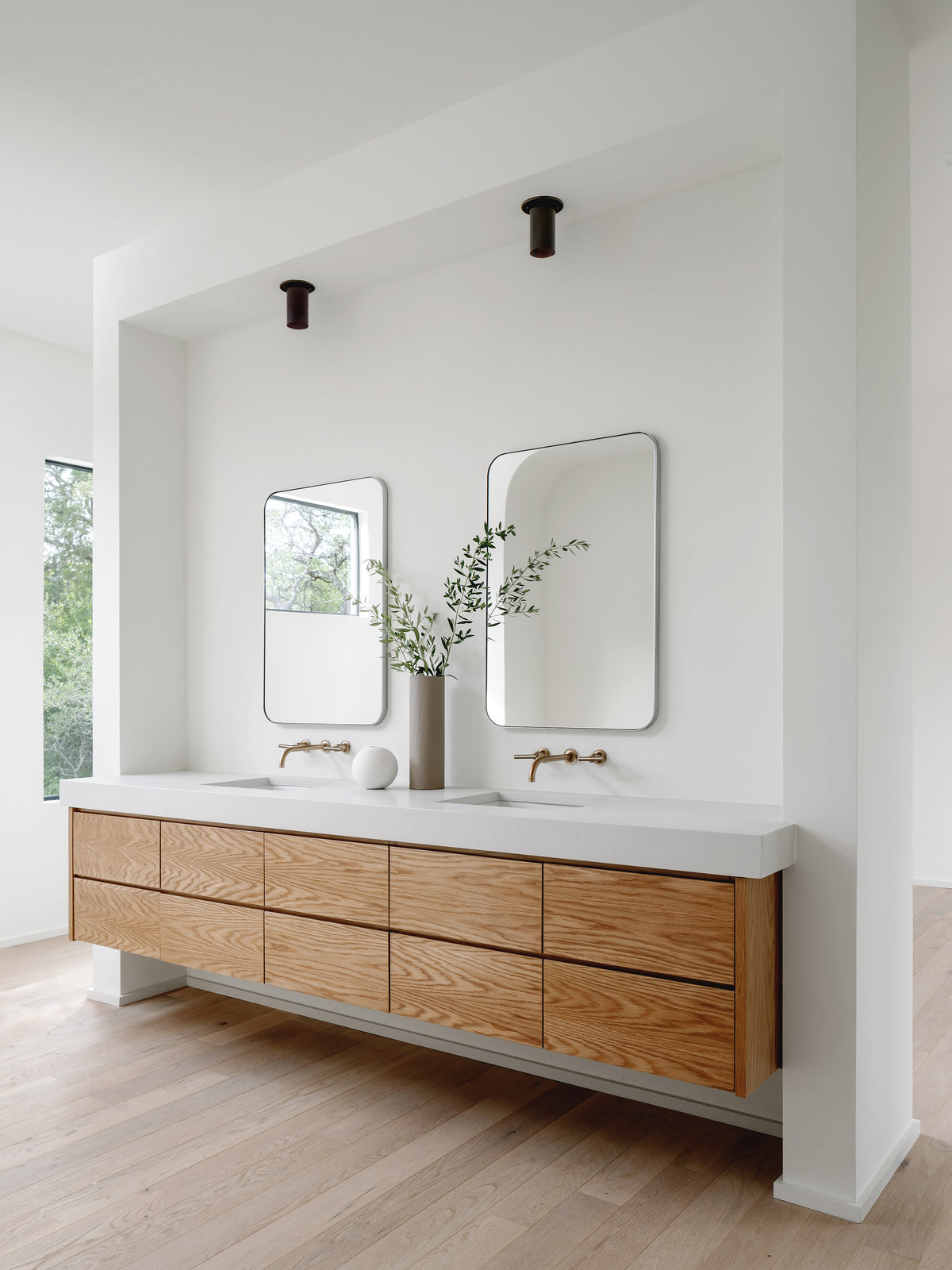Minimalist bathroom with a floating wooden vanity, dual rectangular mirrors, and wall-mounted brass faucets. Cove Flush Spot fixtures by Long Made Co. add subtle illumination, enhancing the natural light from the nearby window.
