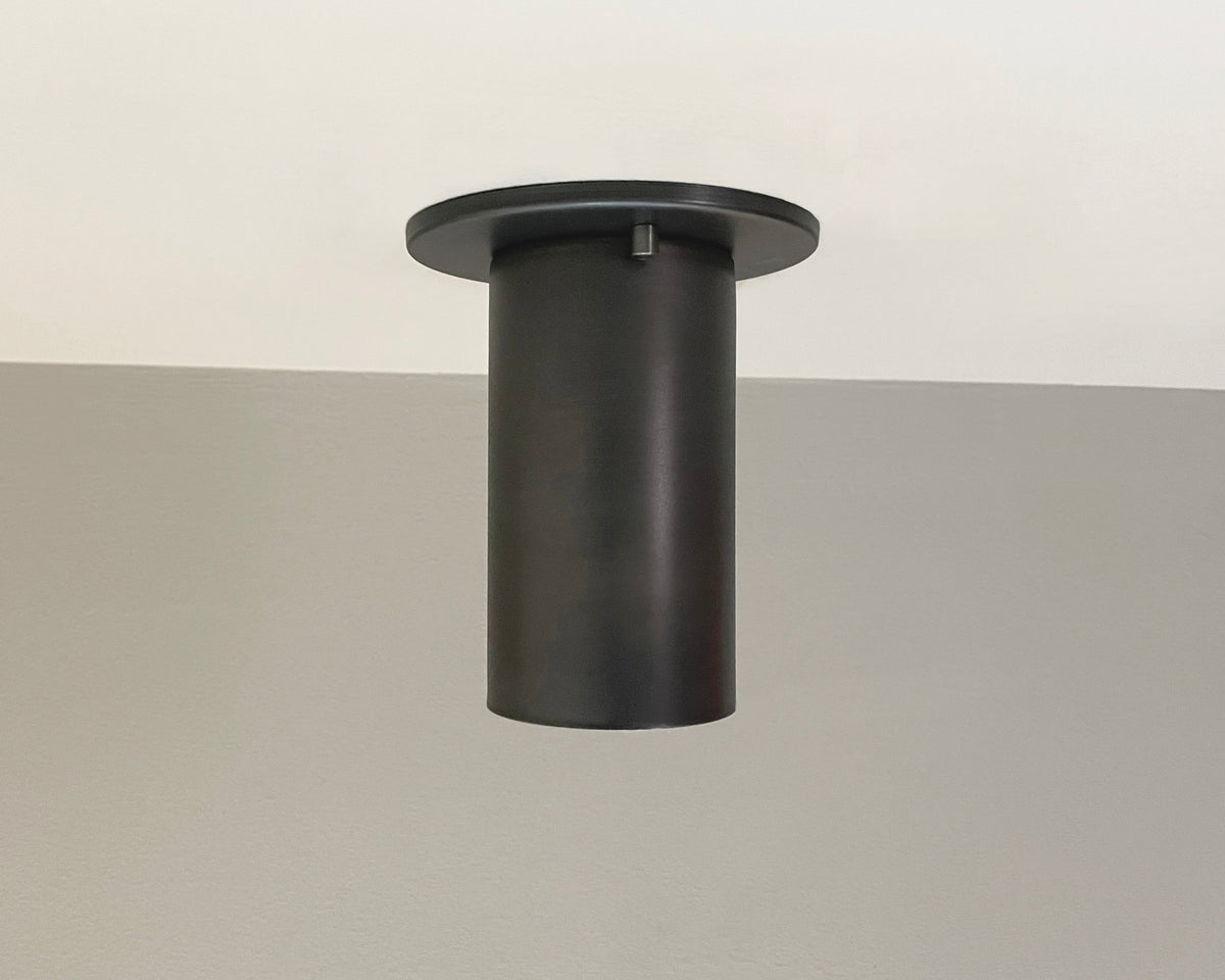 The Cove Flush Spot by Long Made Co., a black solid steel downlight from the Cove Series, is installed on a white and beige ceiling, featuring a sleek, modern design inspired by minimalist spotlights.