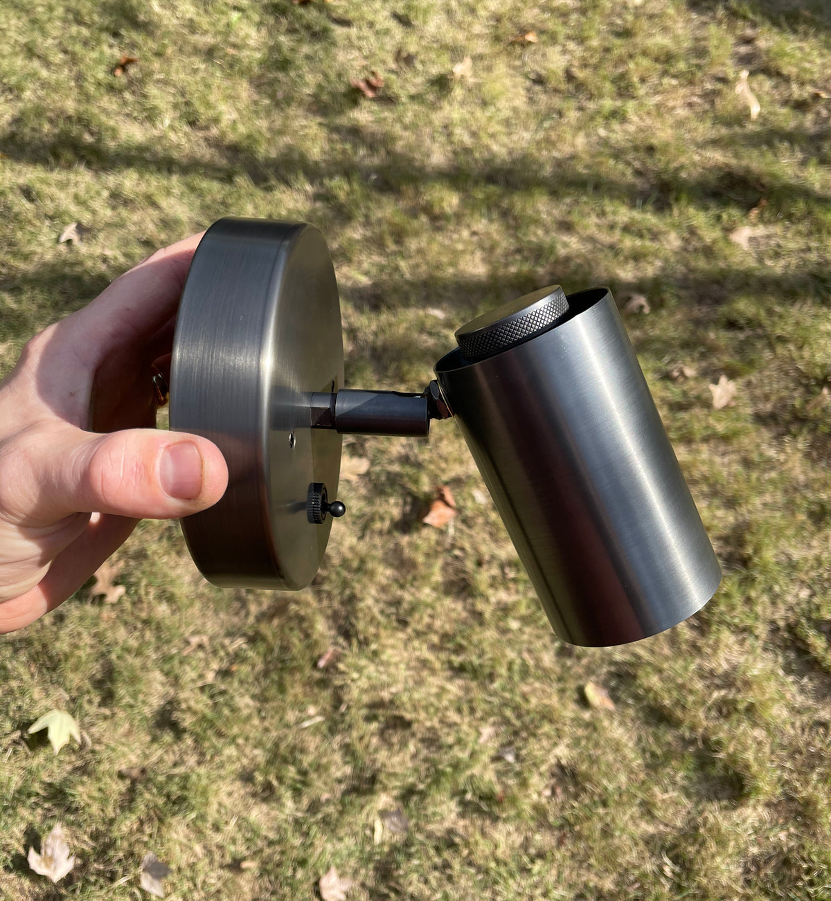 A hand holds the Long Made Co. Brass Cylinder Wall Lamp—an adjustable, wall-mounted light with a brushed metal finish, shown outdoors among dry grass and leaves. It's a perfect mid-century modern fixture to illuminate any space.