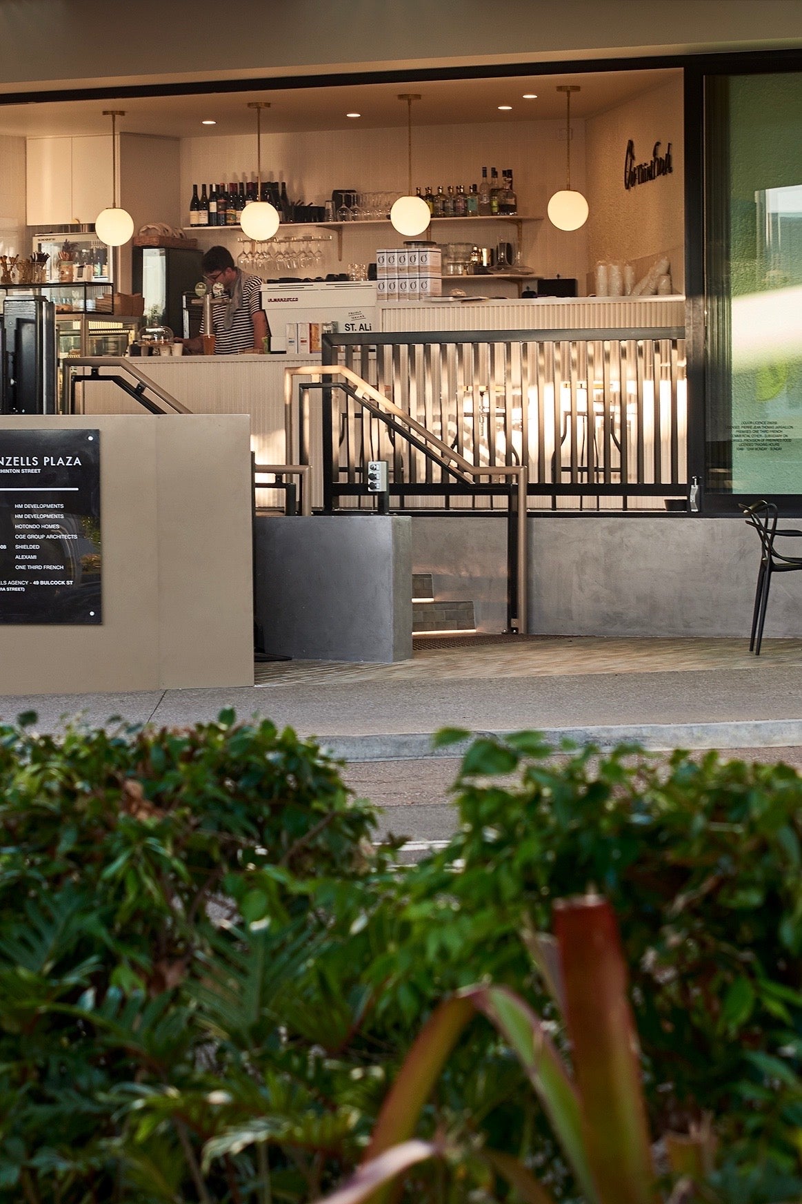 A modern café with large windows features Long Made Co.'s Sphere Solo Drop - 10" lights above the counter, where a barista works. Plants and a sign are visible in the foreground, with seating inside the café.