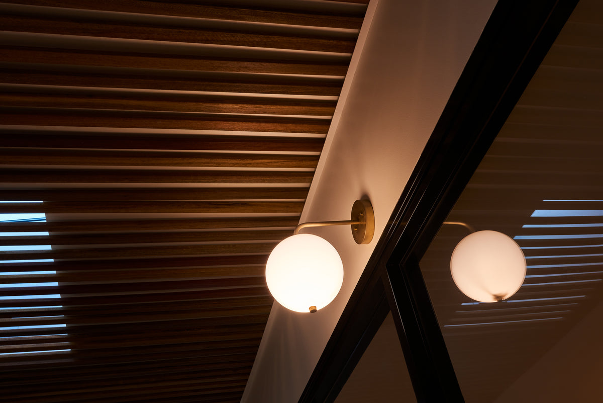 A Wade Wall 8" Globe by Long Made Co., featuring a handmade brass base and acrylic globe shade, is mounted by the window, casting soft light. Overhead, wooden ceiling slats filter gentle rays through their gaps.