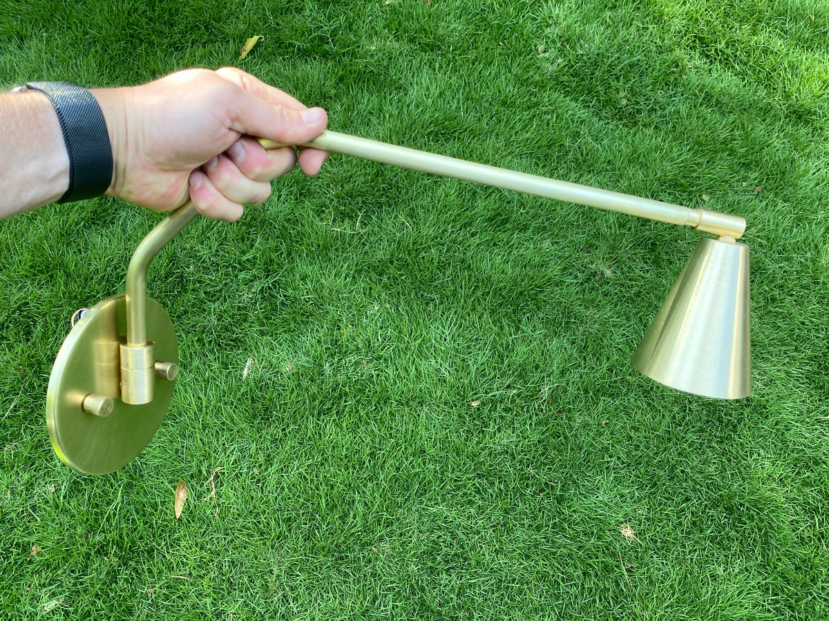 A hand holds the Long Made Co. Tiel Arch Wall Lamp, a handmade brass sconce with an adjustable arm and cone shade, highlighting its modern design against a green grass backdrop.