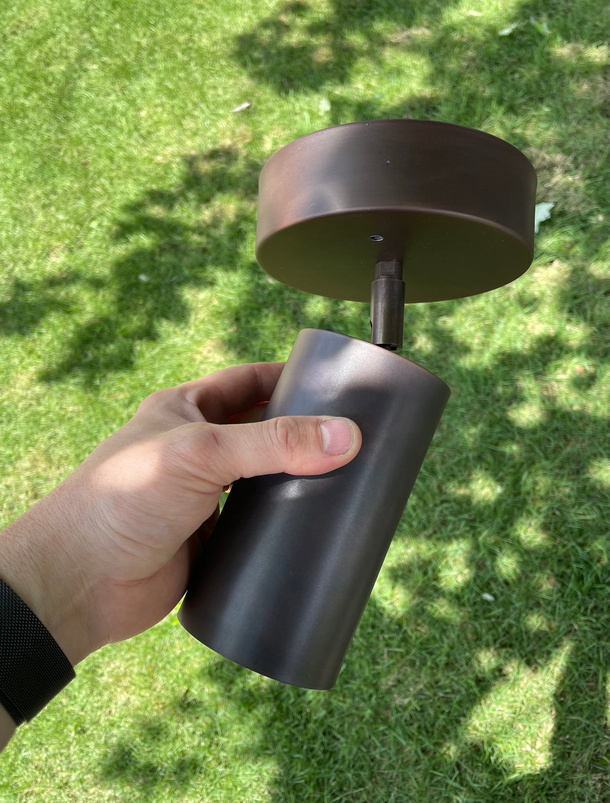 A hand holds the Long Made Co. Cove Adjustable Chunky Spot, a cylindrical lighting fixture with a bronze finish, over a grassy lawn with sunlight and tree shadows in the background.