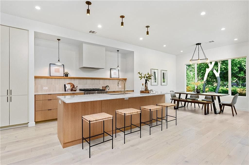 A modern kitchen and dining area features light wood floors, white walls, a large island with four woven barstools, built-in cabinets, large windows overlooking a green yard, and Long Made Co.'s Brass Cylinder Drop Spot pendant lighting.