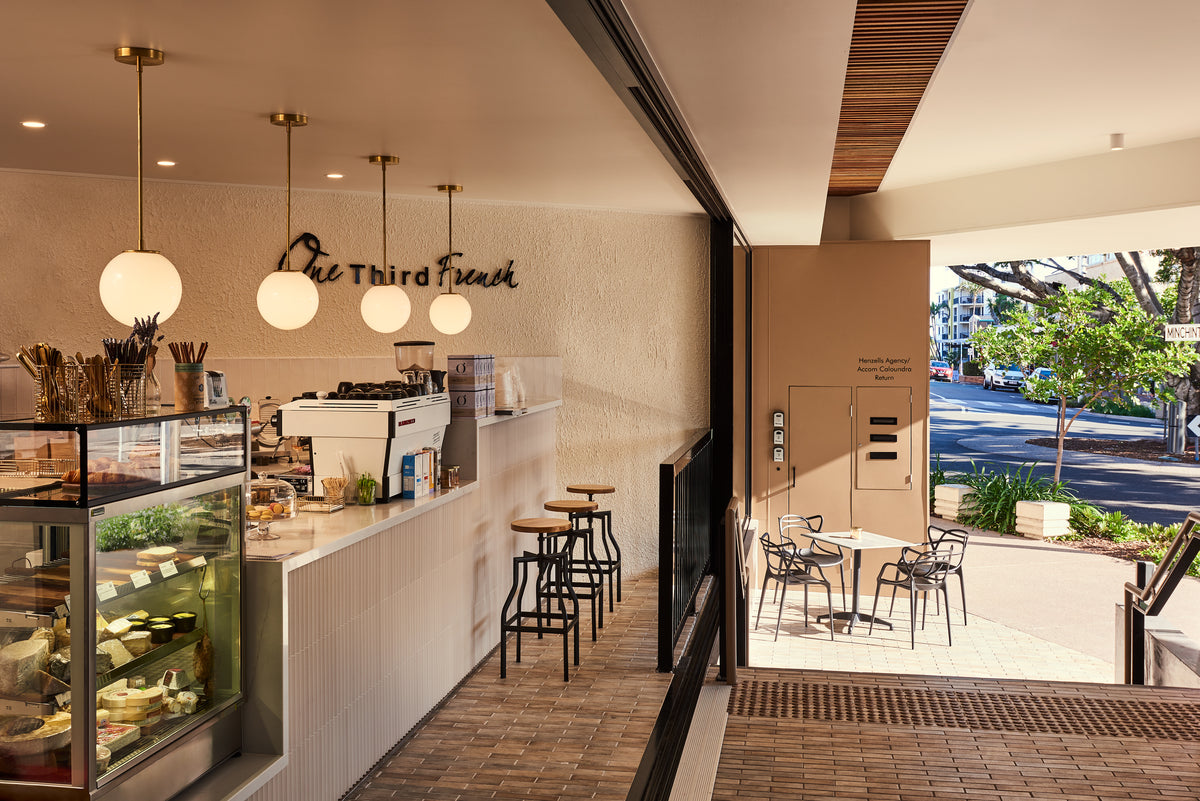 Modern café interior with a white counter, dessert case, stools, and handmade lighting featuring Long Made Co.'s Sphere Solo Drop - 10" solid brass pendant lights. The café opens to outdoor seating overlooking a tree-lined street. Sign: The Third French.