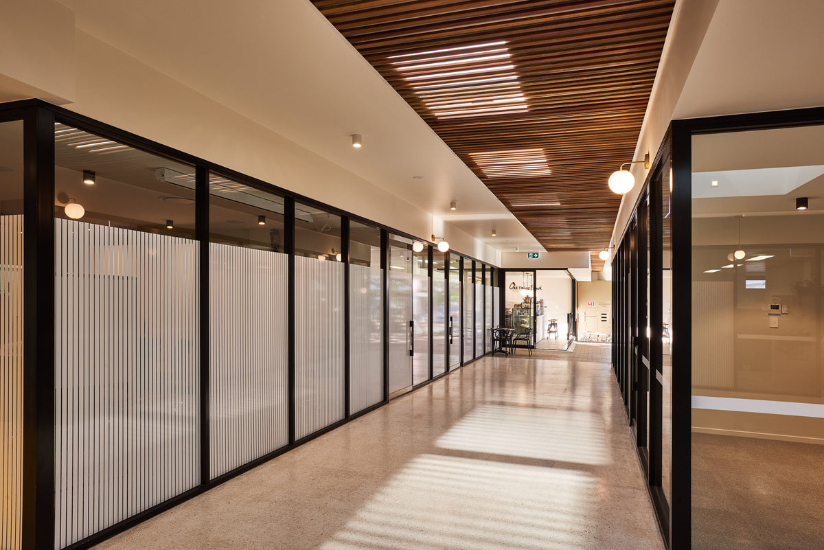 A modern hallway features glass-walled offices, a wood-slat ceiling casting shadows, and bright illumination from Long Made Co.'s Wade Wall 8" Globe lights with round acrylic shades, creating a clean, inviting space.