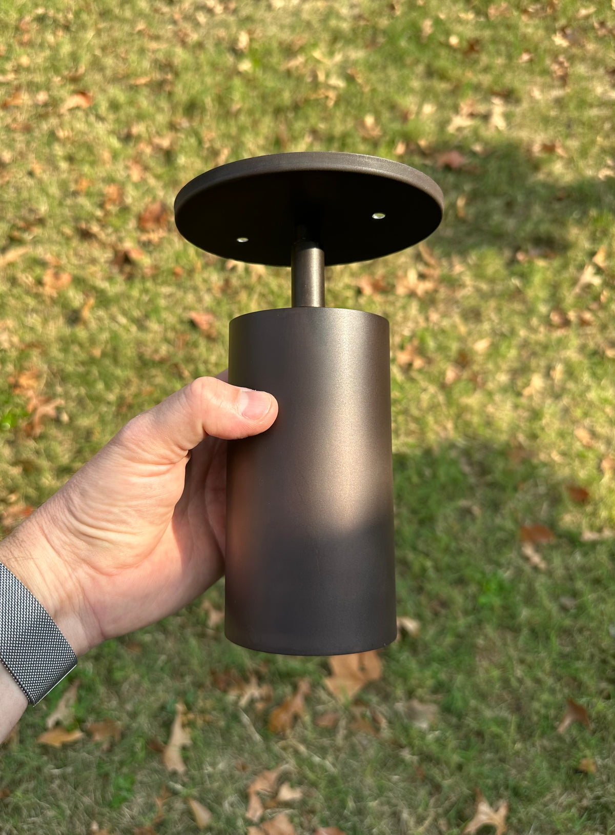 A hand holds the Long Made Co. Cove Adjustable Spot, a cylindrical black solid steel downlight with a round mounting plate, shown outdoors among green grass and scattered brown leaves.