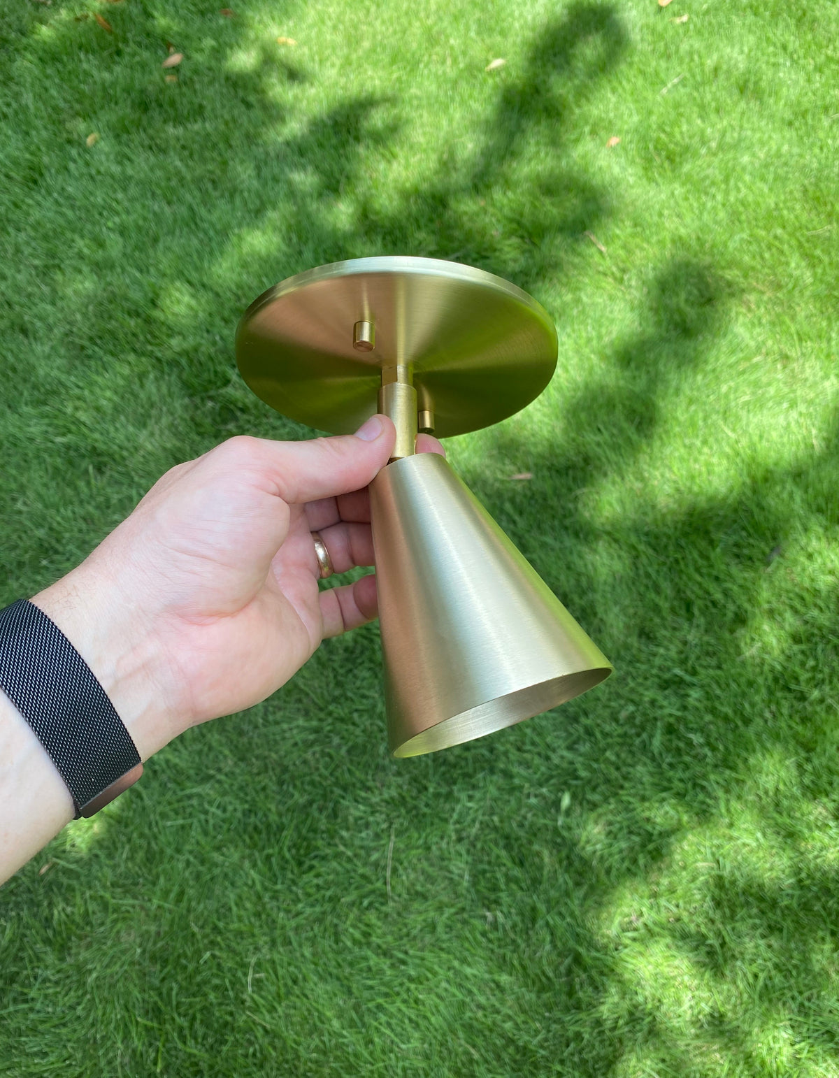 A hand holds the Tiel Wall Spot by Long Made Co., its handmade brass cone gleaming gold in outdoor light against green grass and soft shadows.