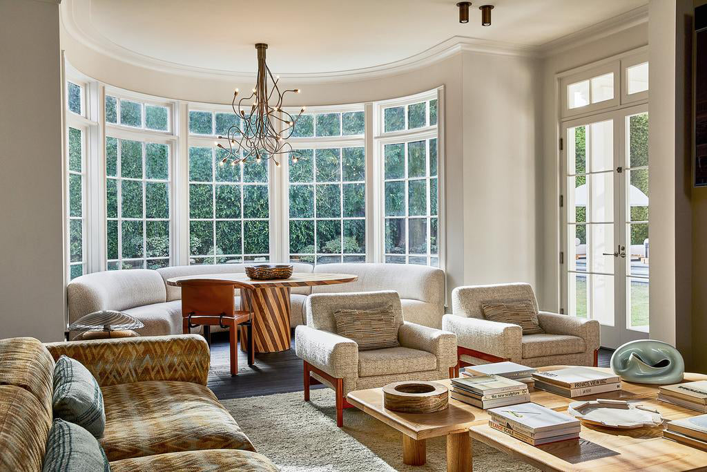 A bright, modern living room with large curved windows, neutral sofas and chairs, a wooden coffee table with books, and a round dining table; Cove Flush Spot by Long Made Co. adds minimalist elegance while greenery is visible outside.