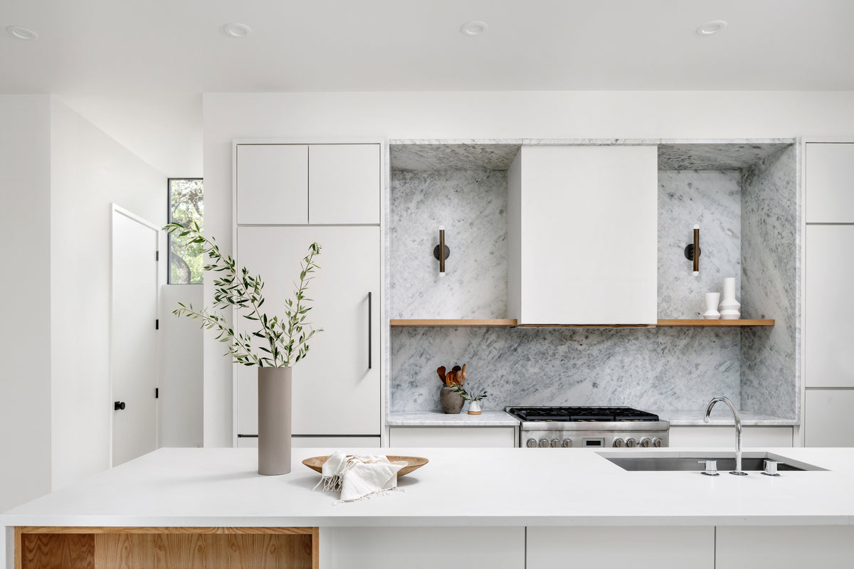 Minimalist modern kitchen with white cabinetry, marble surfaces, a built-in stove, wood accents, neutral decor, a large island topped with a vase of greenery, and the Miro 2 - 12" Sconce by Long Made Co. as a handmade light fixture.