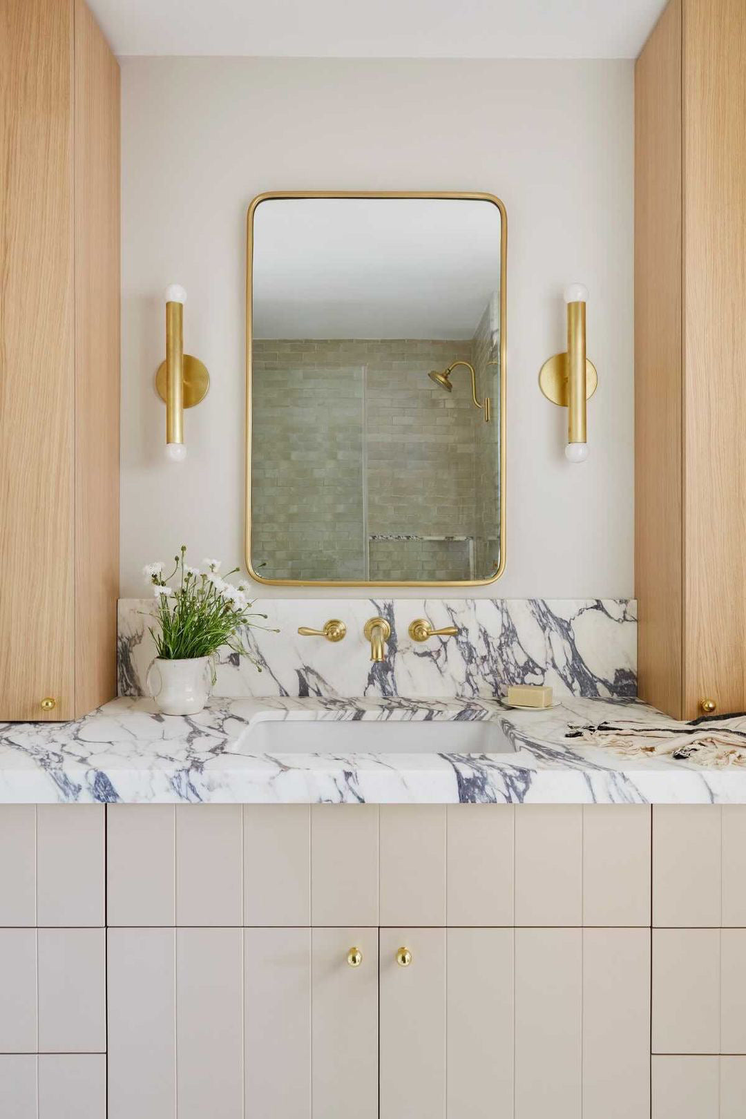 A modern bathroom vanity features a marble countertop, gold fixtures, a rectangular mirror with a gold frame, and light wood cabinets. Beside the sink is a potted plant and the Miro 2 - 12" Sconce by Long Made Co. brass wall lighting.