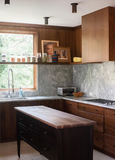 A modern kitchen features dark wood cabinets, a marble backsplash, and a small island. Long Made Co.’s Cove Flush Spot provides task lighting above framed photos, a toaster oven, and dishes as natural light streams in.