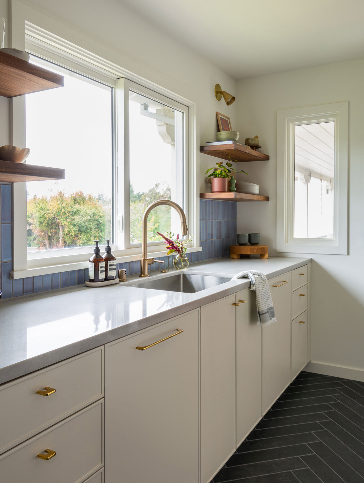 Bright modern kitchen with white cabinets, gold hardware, a large sink under the window, blue tile backsplash, wooden shelves with plants and dishes, chevron-patterned dark floor tiles, and Long Made Co.'s Tiel Wall Spot lighting fixture.