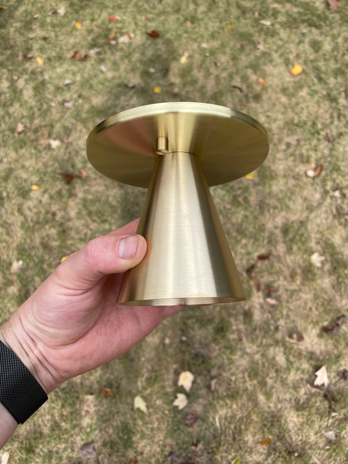 A hand holds the Tiel Flush Spot by Long Made Co., a small, gold-colored, cone-shaped solid brass fixture with a flat circular top—an elegant alternative to recessed can lights—shown outdoors on leafy grass.