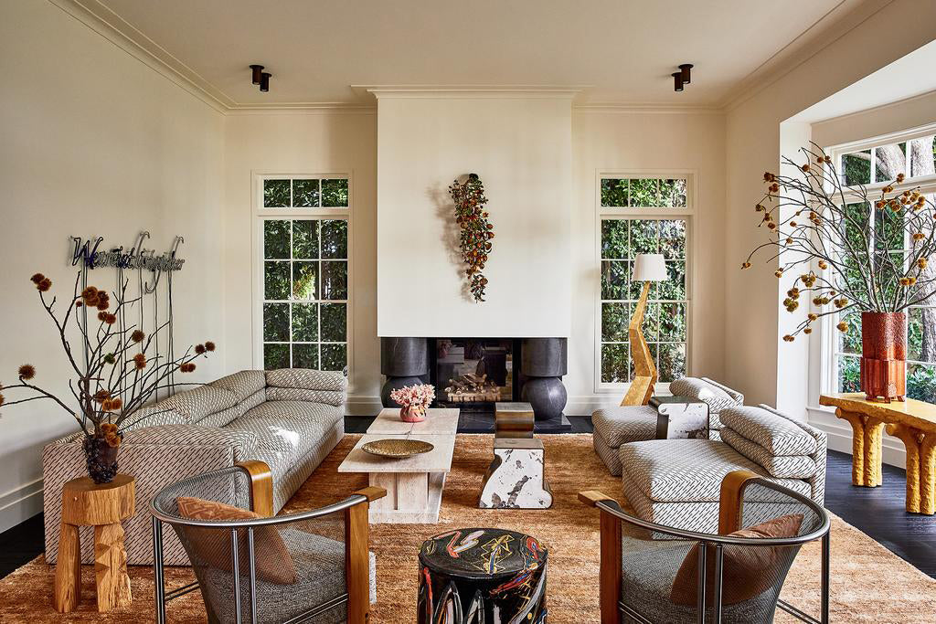 A stylish living room with neutral sofas, modern armchairs, a fireplace, wall art, tall windows, and plants. Cove Flush Spot lights by Long Made Co. offer minimalist task lighting that highlights the patterned furniture and wood coffee table.