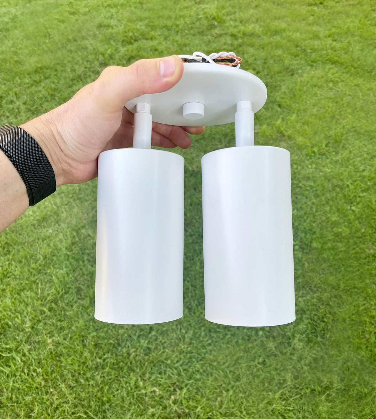 A hand holds the Long Made Co. Cove Double / Triple Spot, a white dual-cylinder directional ceiling downlight with exposed wires, set against green grass.